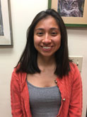 A young tan woman with mid-length dark hair wears a gray tank-top and coral-colored jacket and smiles in front of a hallway of framed photos.